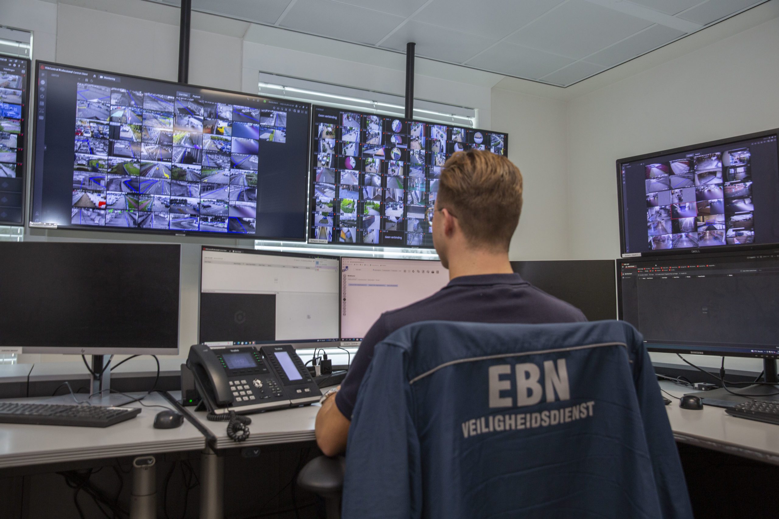 Centralist in EBN-jas kijkt in de meldkamer naar een wand met camerabeelden